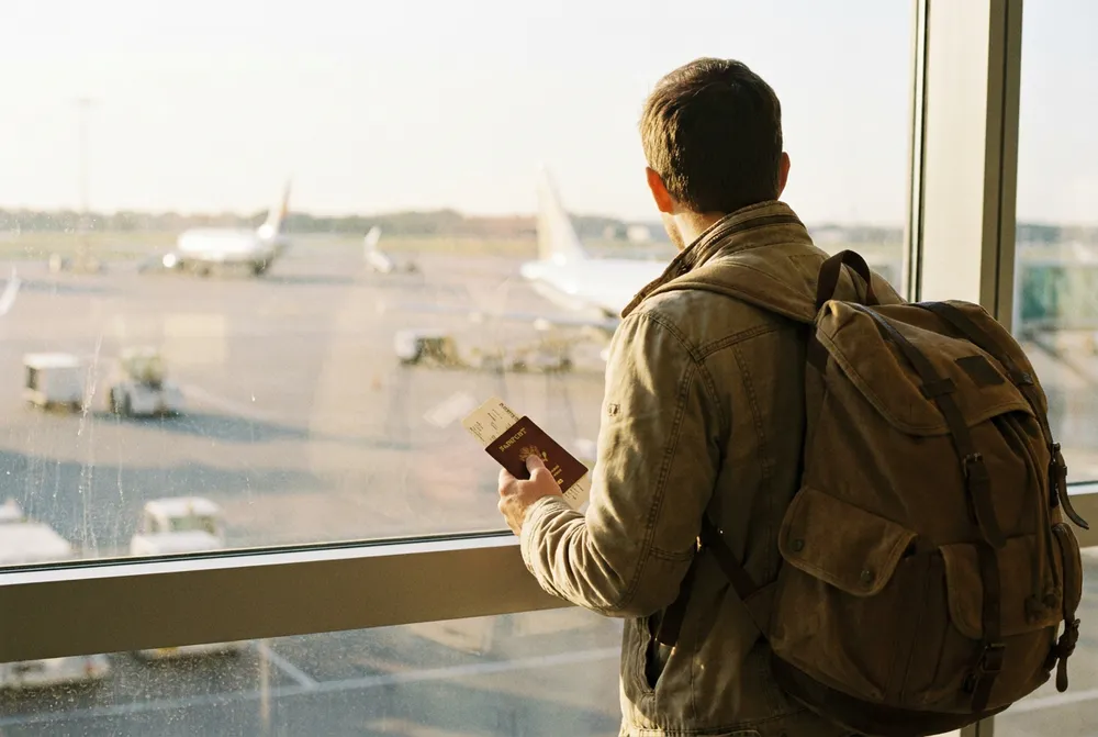 Persona con mochila y pasaporte en mano mirando la pista por la ventana del aeropuerto